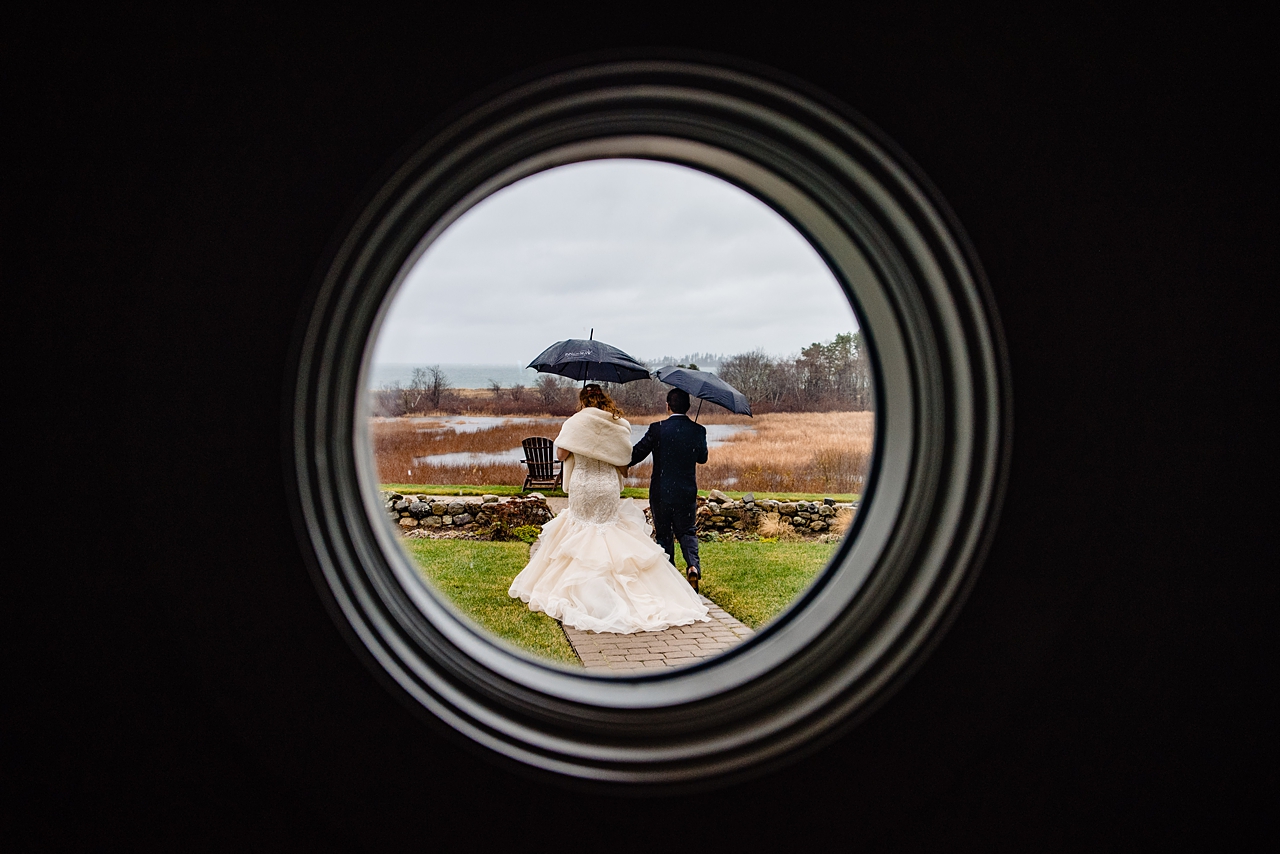Wedding couple with umbrellas walking away and photographed through a round window.